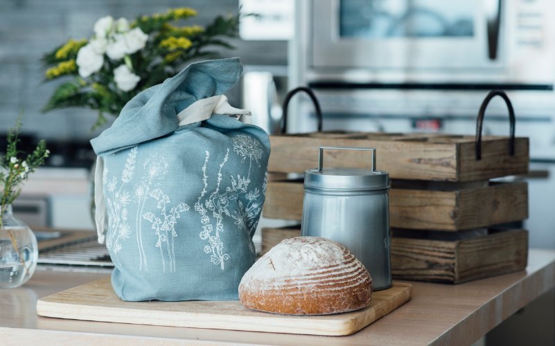 Linen bread bag, hand printed in white floral design onto duck egg blue linen. The bread bag is on a wooden bread board with a loaf of sourdough