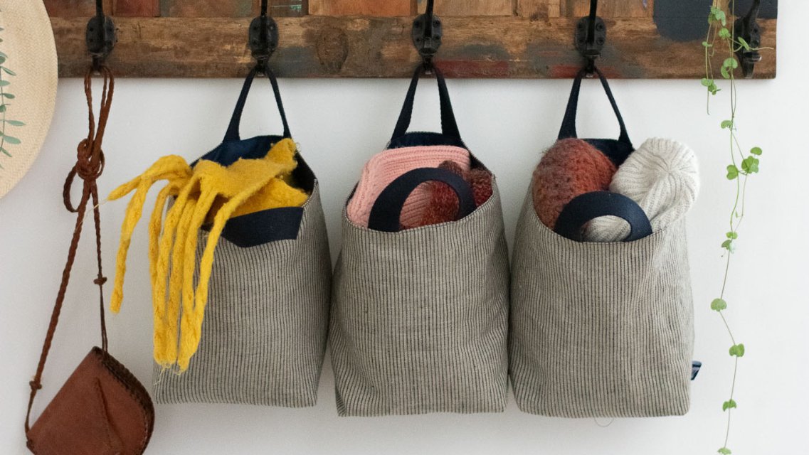 Three Large Fabric Storage Pots with Handles handing on a wooden coat rack with wrought iron handles.  Made from Striped blue and white linen and filled with hats, gloves and scarves.