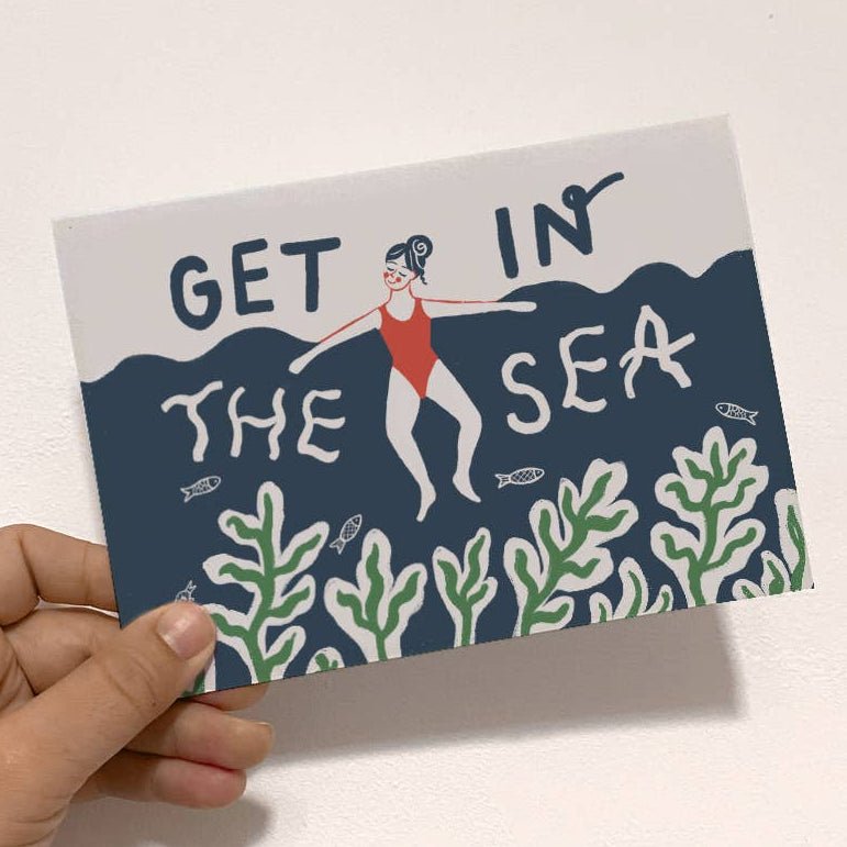 Hand holding a postcard with 'Get in the Sea' text and sea-themed illustrations sold by Helen Round