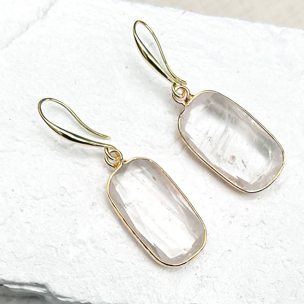 A pair of Quartz Rectangle Drop Earrings with rectangular clear stone pendants, displayed on textured white stone slabs. Sold by Helen Round