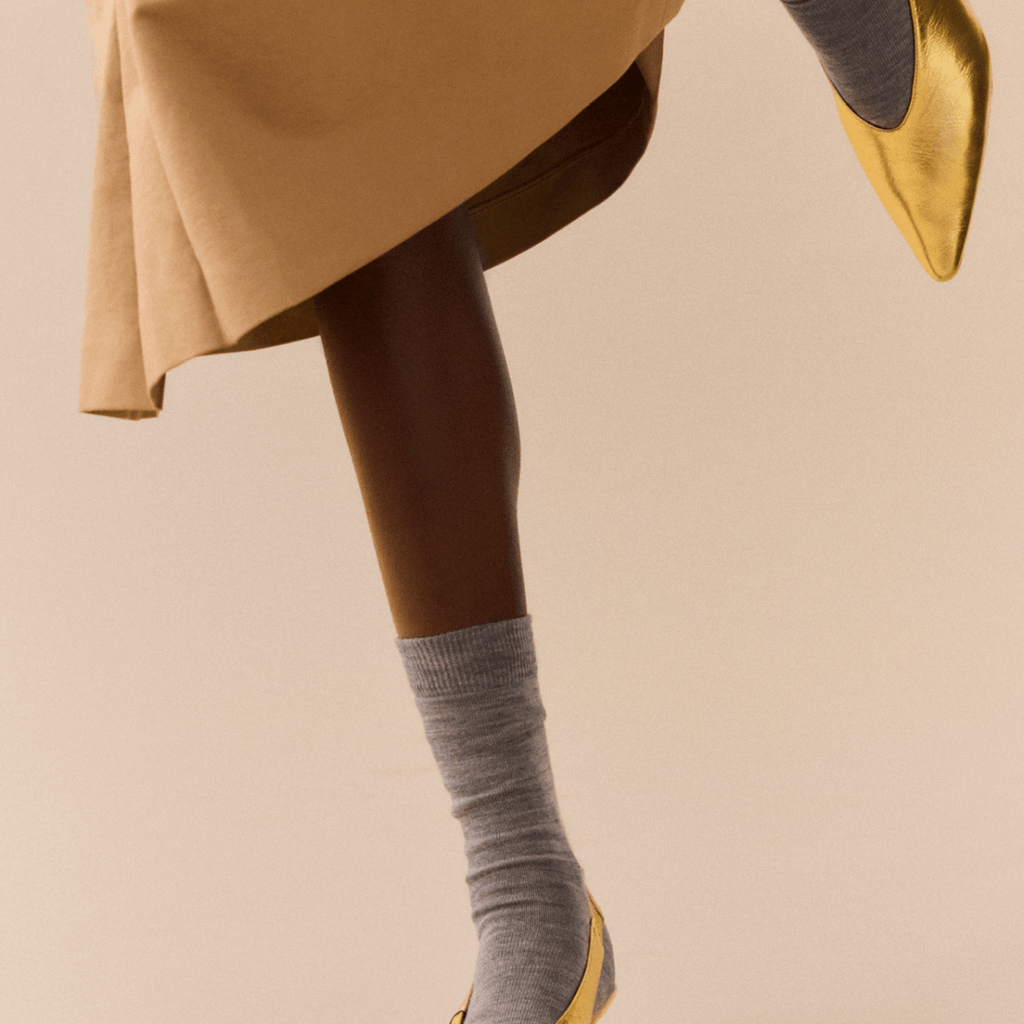 Person wearing gold shoes and grey merino socks on a beige background