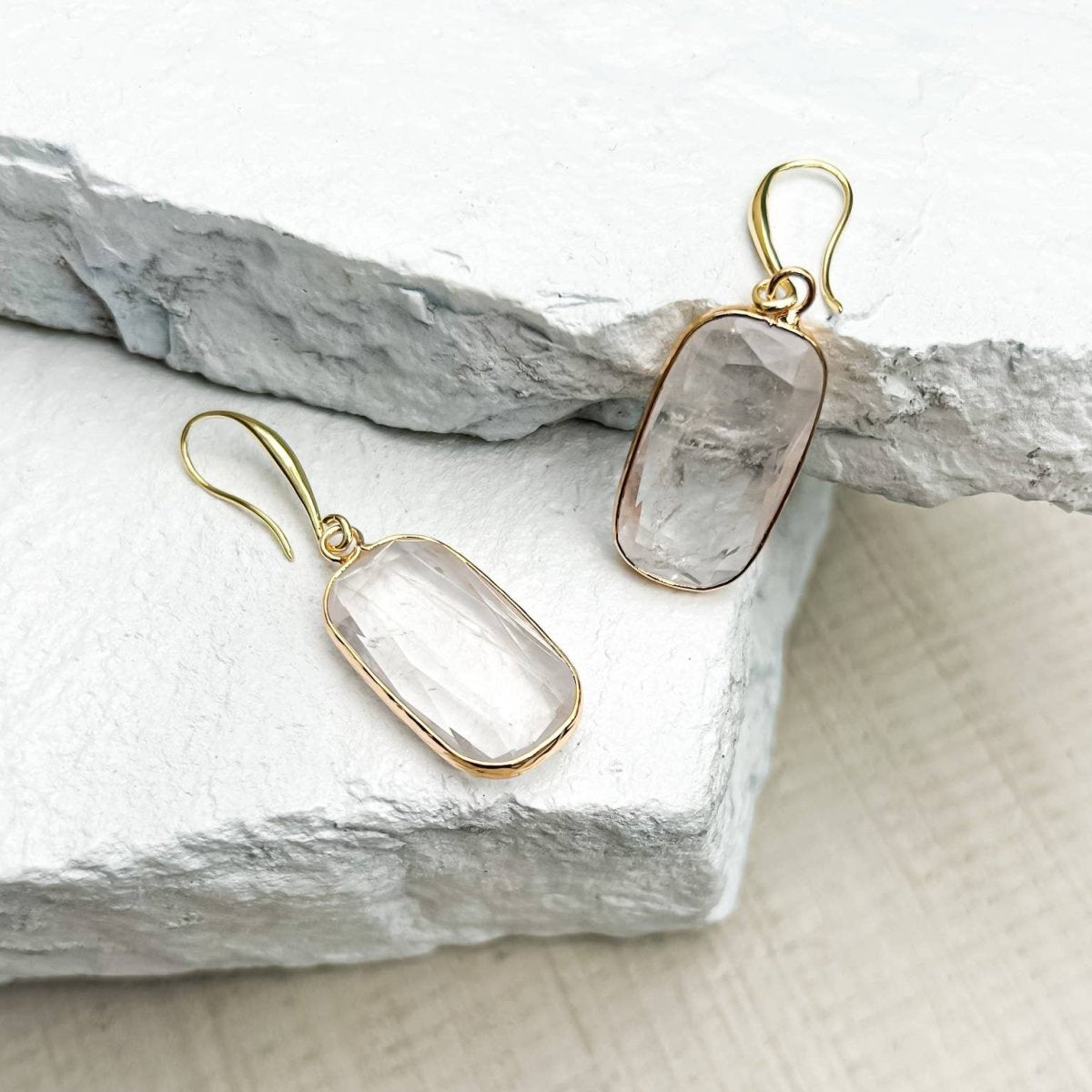 A pair of Quartz Rectangle Drop Earrings with rectangular clear stone pendants, displayed on textured white stone slabs.
