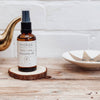 Bottle of moksa daily calm cleansing oil on a wooden coaster with a teapot and bowl in the background.