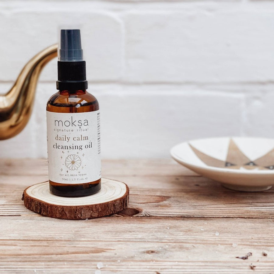 Bottle of moksa daily calm cleansing oil on a wooden coaster with a teapot and bowl in the background.