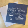 Navy blue card with white illustrations of a coastal scene on a wooden surface
