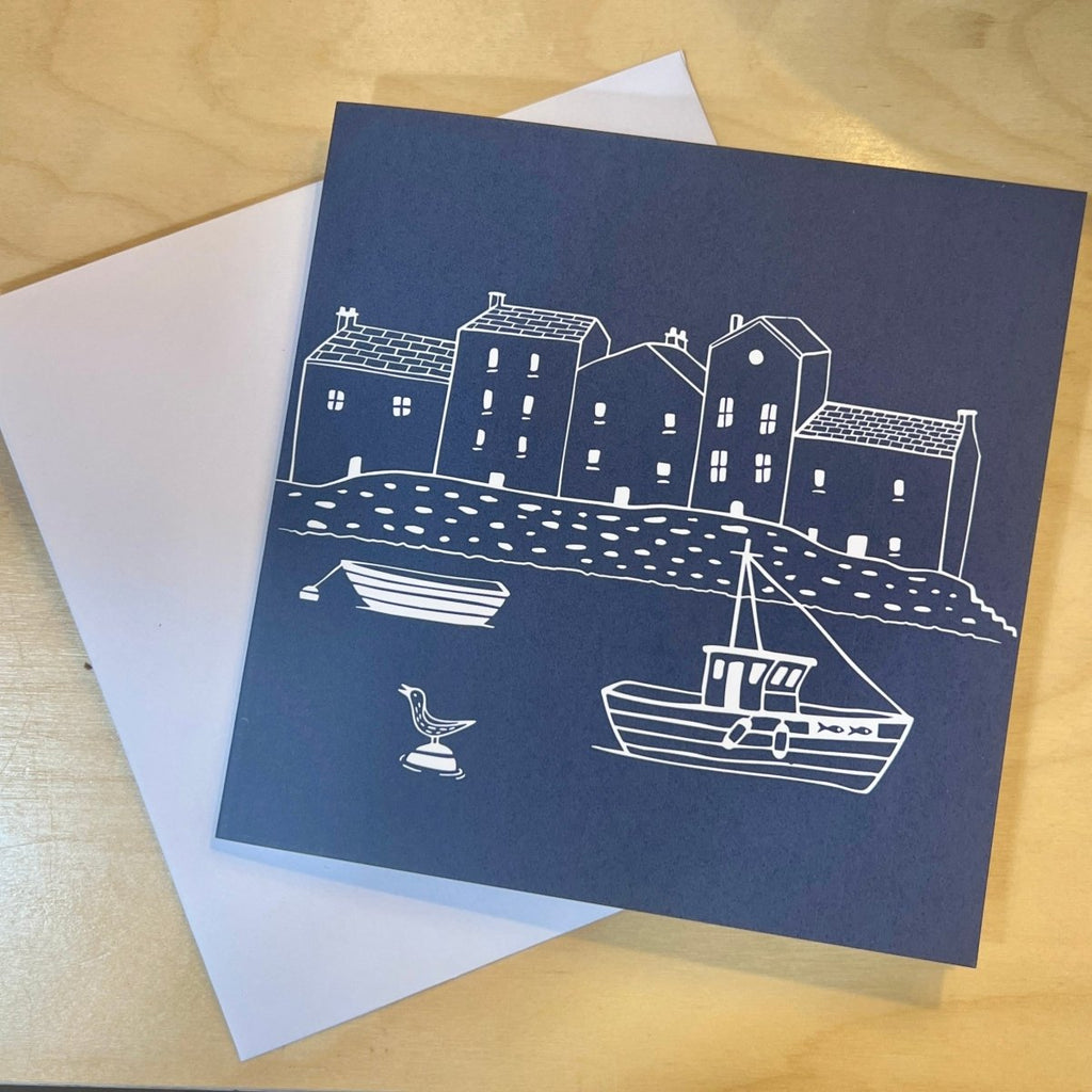 Navy blue card with white illustrations of a coastal scene on a wooden surface