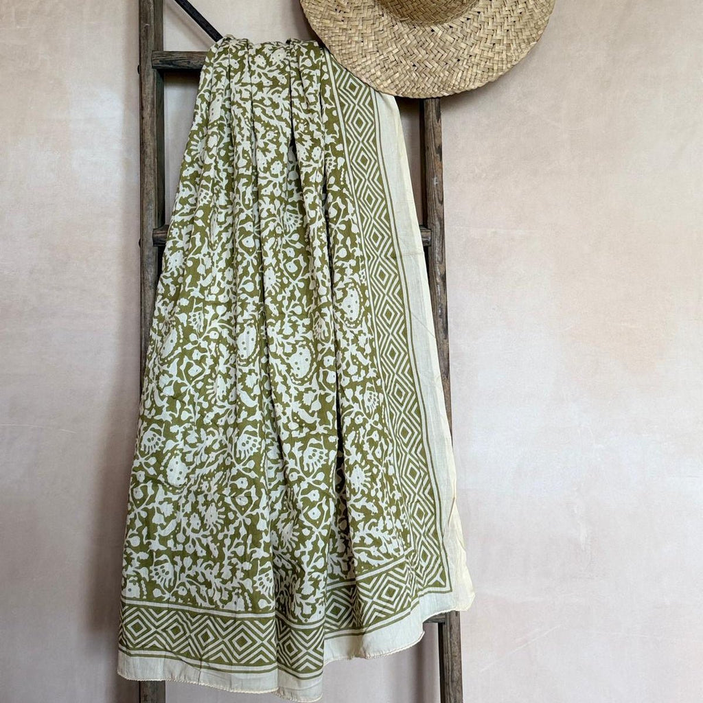 Green patterned sarong and straw hat on a wooden ladder against a beige wall.