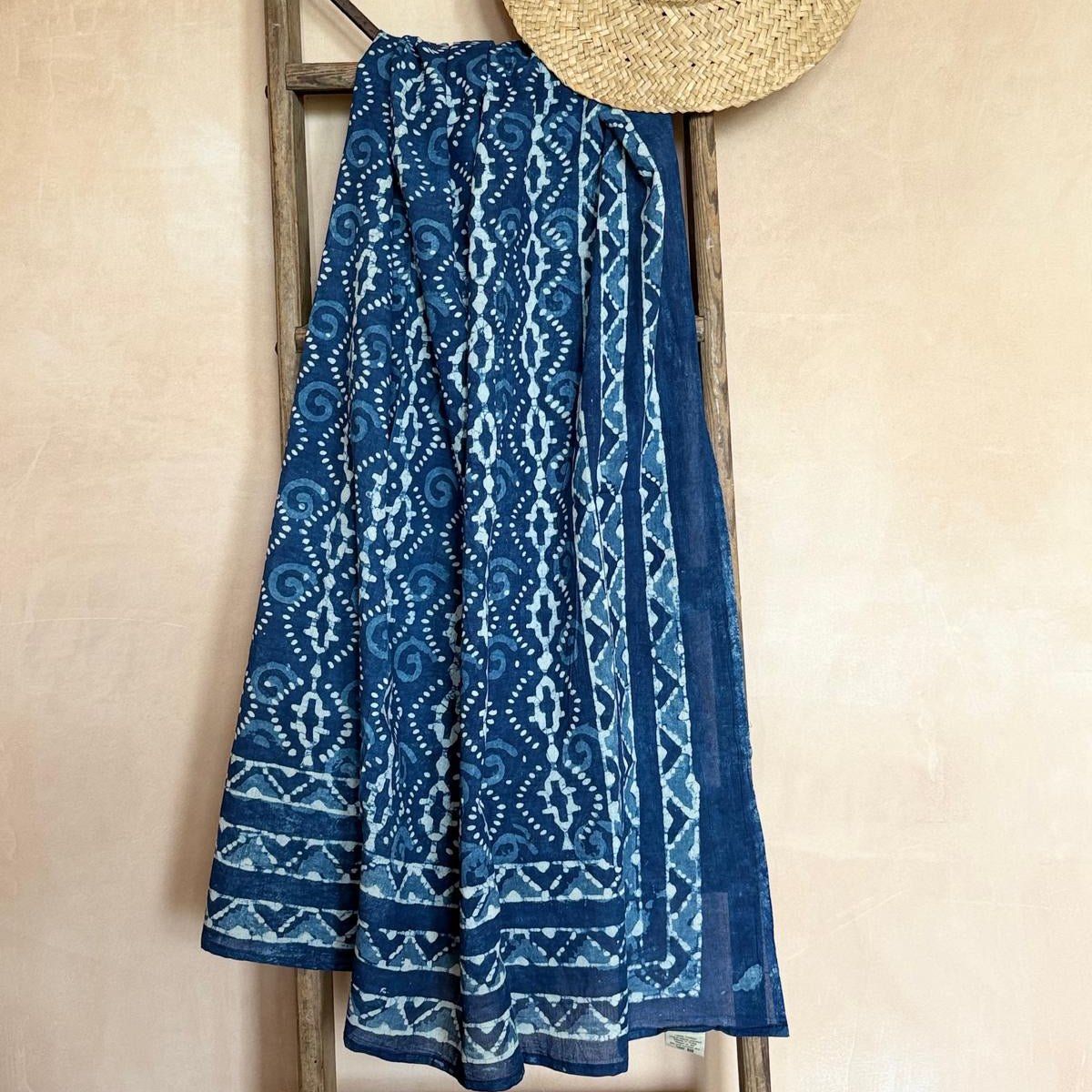 Blue patterned sarong and straw hat on a wooden ladder against a beige wall