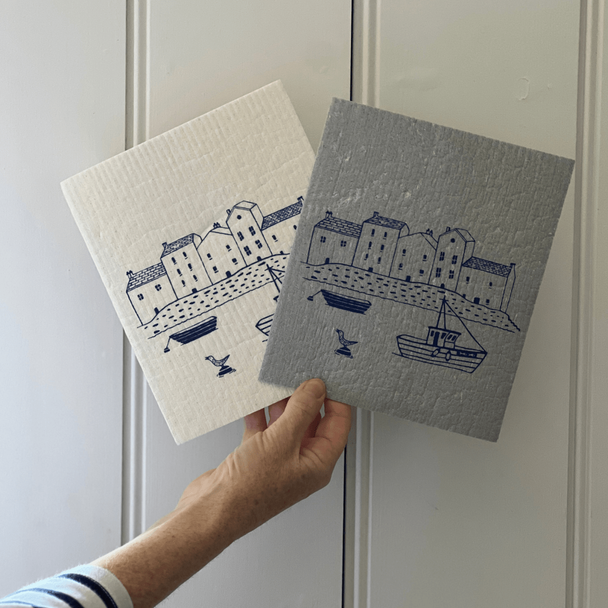 Two dishcloths with blue illustrations of houses and boats held against a white background.