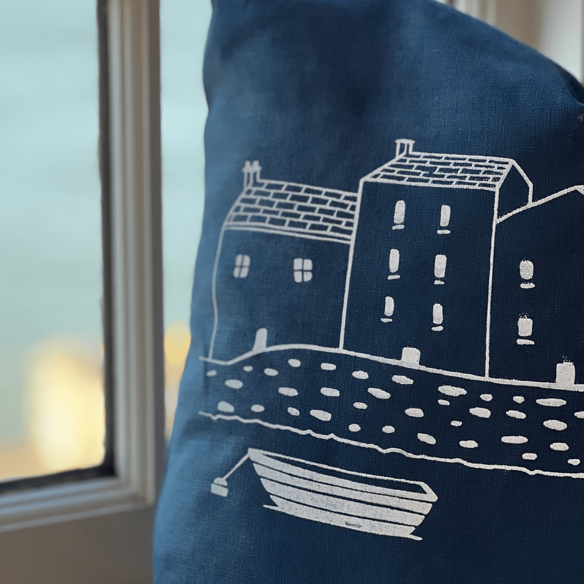 Navy blue pillow with white house and boat design against a blurred indoor background