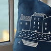 Navy blue pillow with white house and boat design against a blurred indoor background