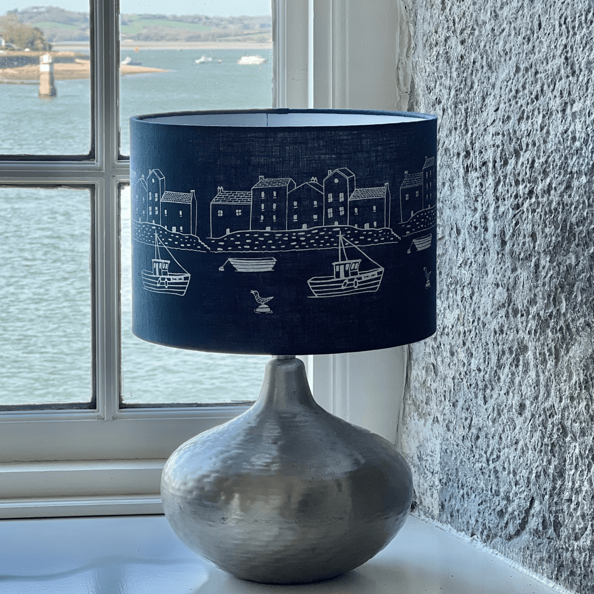 Lamp with a navy blue linen shade featuring white illustrations, placed on a windowsill with a view of water and land.