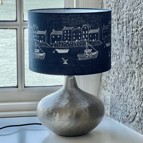 Lamp with a linen navy blue lampshade featuring white illustrations of houses and boats, placed on a surface near a window from a collection by Helen Round.