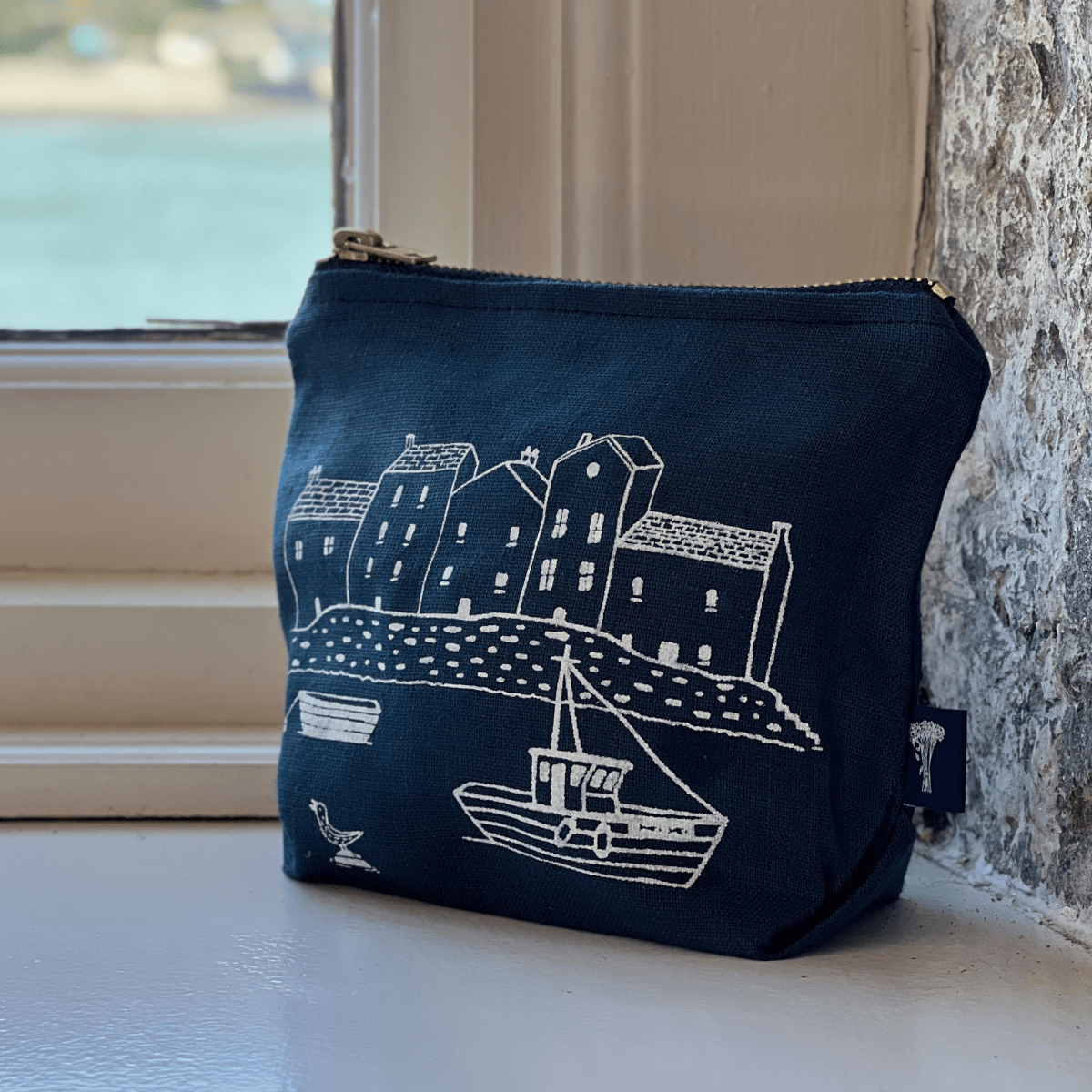 Navy blue linen makeup bag with white boat and building design on a windowsill.