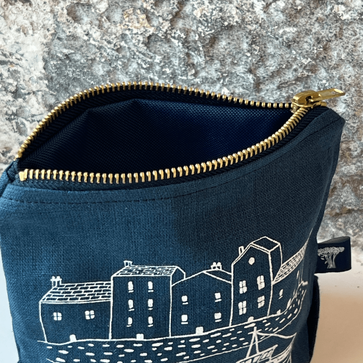 Blue linen makeup bag with white house illustration and gold zipper on a stone surface from Helen Round.