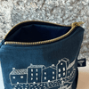 Blue linen makeup bag with white house illustration and gold zipper on a stone surface from Helen Round.