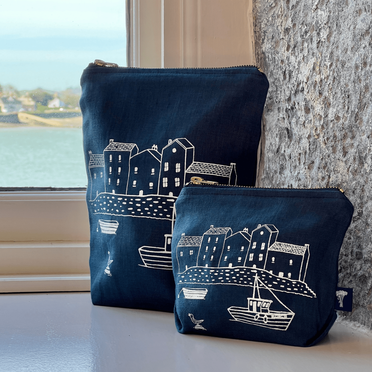 Two navy blue linen large for toiletries and small for makeup with white illustrations of houses and boats on a windowsill from Helen Round.