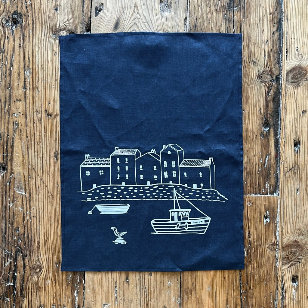 Navy blue tea towel with white printed design of a coastal scene on a wooden surface by Helen Round 