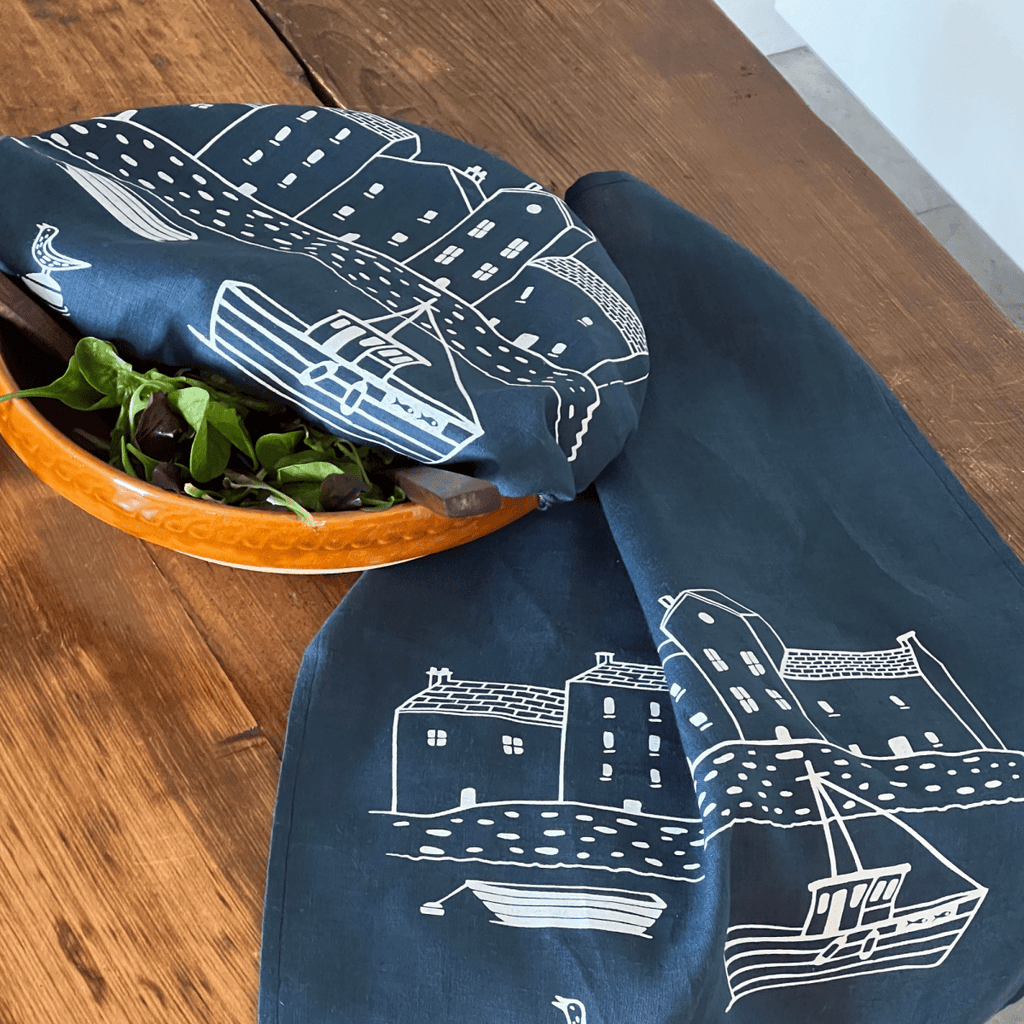 Navy blue tea towel with white boat and house pattern draped over a wooden surface with a bowl of salad covered with a matching navy blue linen bowl cover.