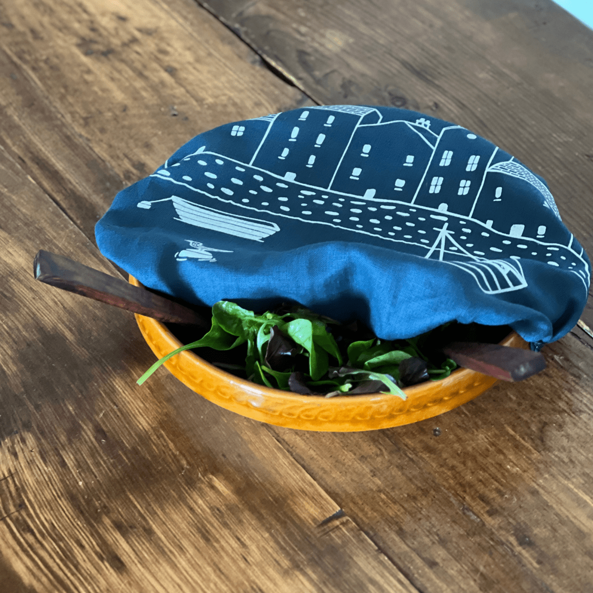 Bowl of salad with a navy blue linen bowl cover featuring white house illustrations on a wooden surface from Helen Round.