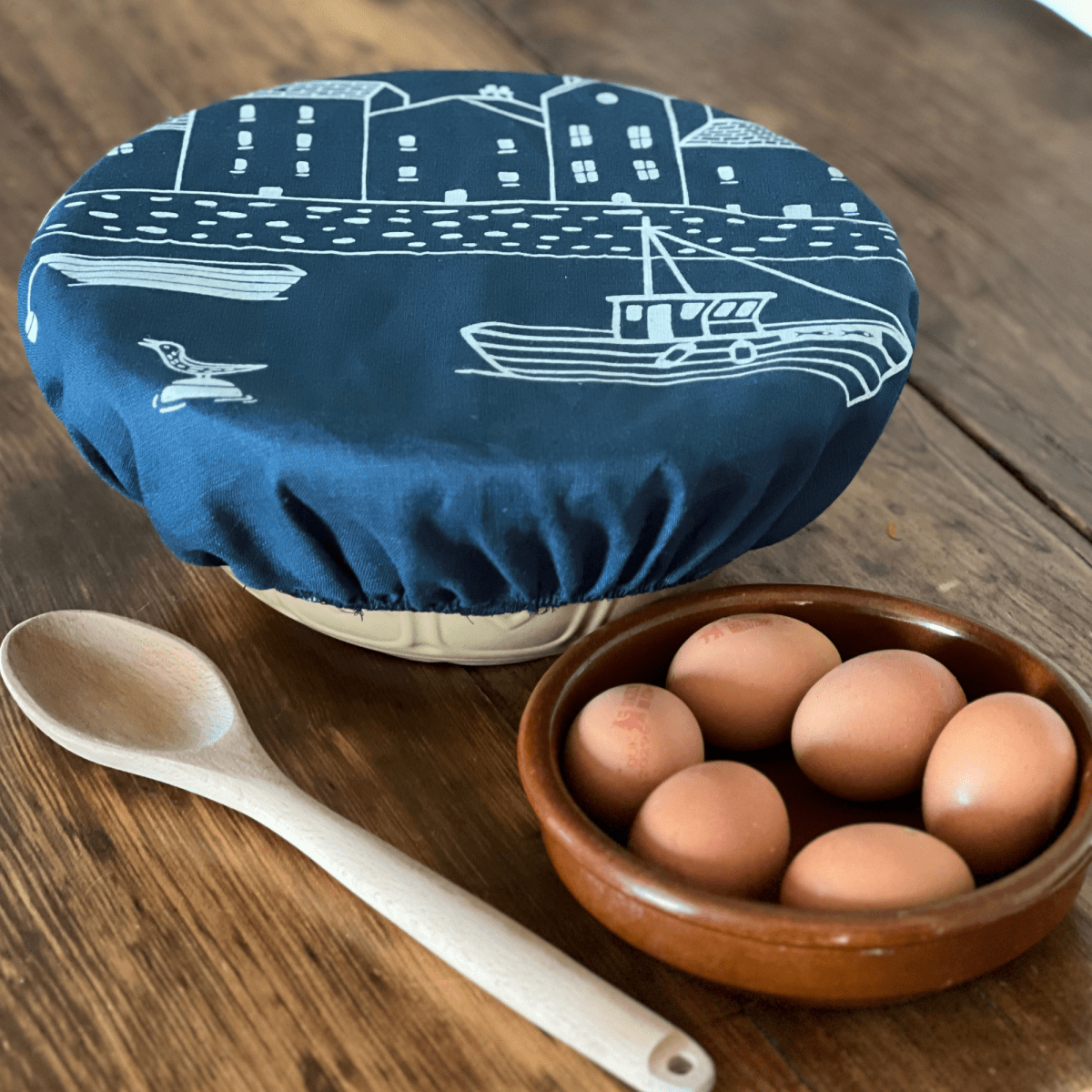 Large navy blue linen elasticated bowl cover with white boat design, wooden spoon, and brown bowl with eggs on a wooden surface from Helen Round.