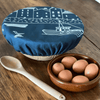 Large navy blue linen elasticated bowl cover with white boat design, wooden spoon, and brown bowl with eggs on a wooden surface from Helen Round.
