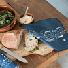 Sandwich on a wooden board with bread, salad, and butter, next to a blue linen reusable sandwich wrap with white designs on a wooden table from Helen Round