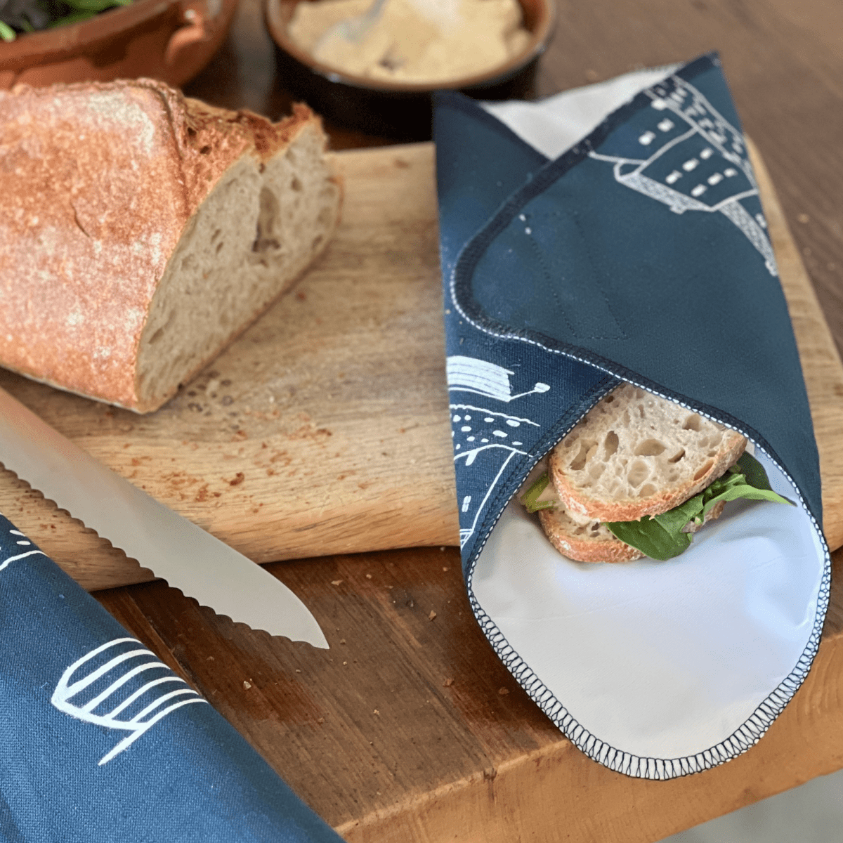 Sandwich wrapped in a blue reusable wrap on a wooden cutting board with bread and a knife.