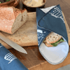 Sandwich wrapped in a blue reusable wrap on a wooden cutting board with bread and a knife.
