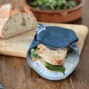 Sandwich wrapped in a blue linen sandwich wrap on a wooden table with bread and salad in the background