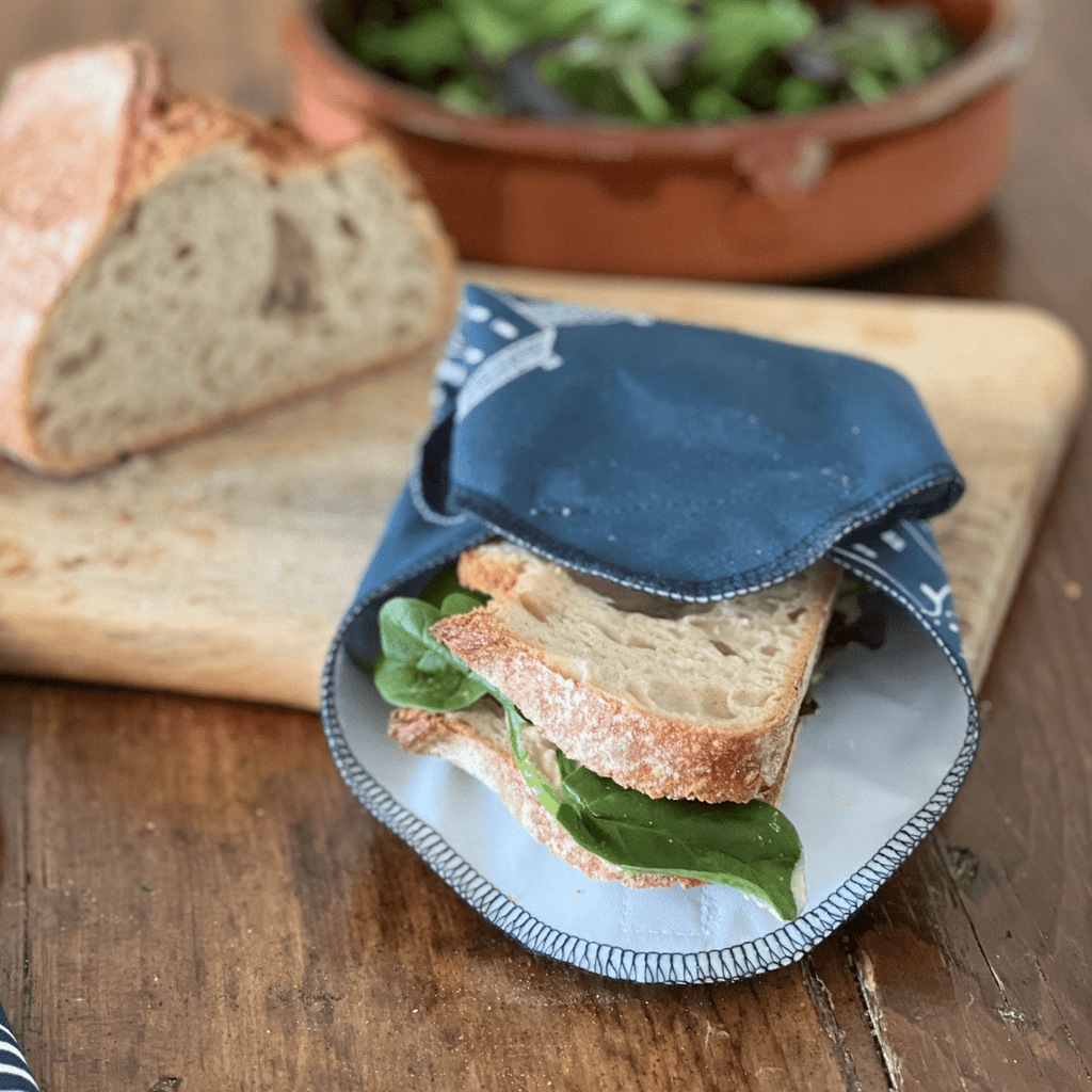Sandwich wrapped in a blue linen sandwich wrap on a wooden table with bread and salad in the background