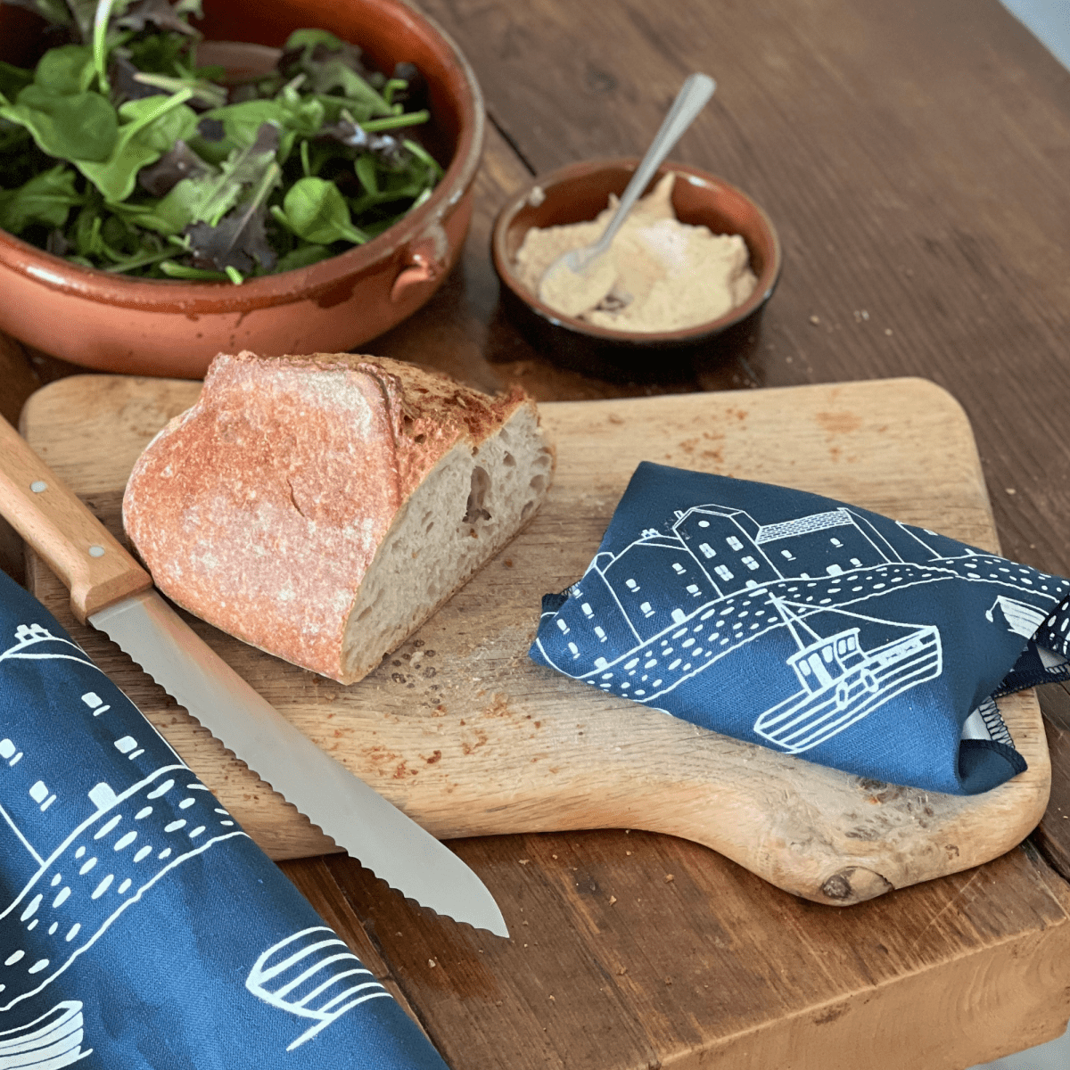 Loaf of bread on a wooden cutting board with a knife, wrapped reusable linen sandwich wrap, and salad in the background.