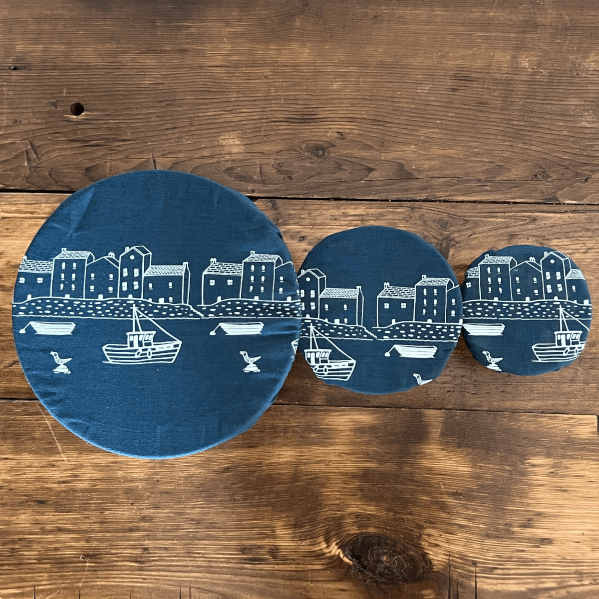 Three blue linen bowl covers with white illustrations of a coastal scene on a wooden surface from Helen Round.