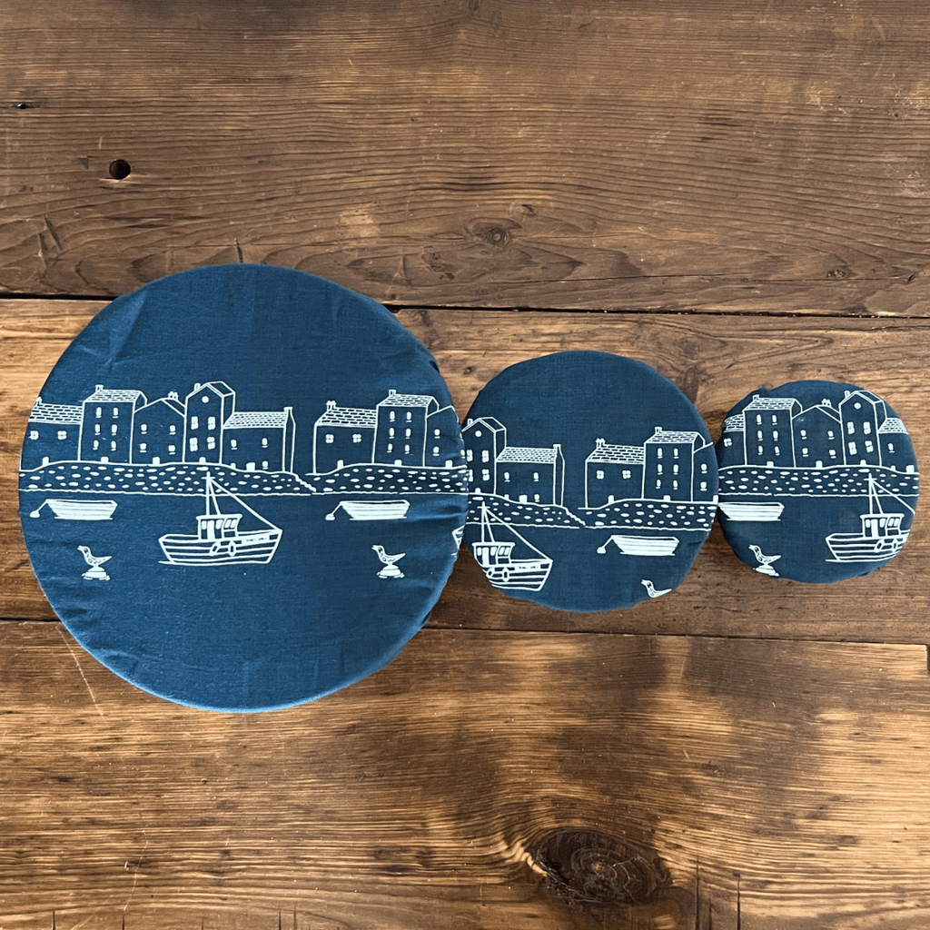Three blue linen bowl covers with white illustrations of a coastal scene on a wooden surface from Helen Round.