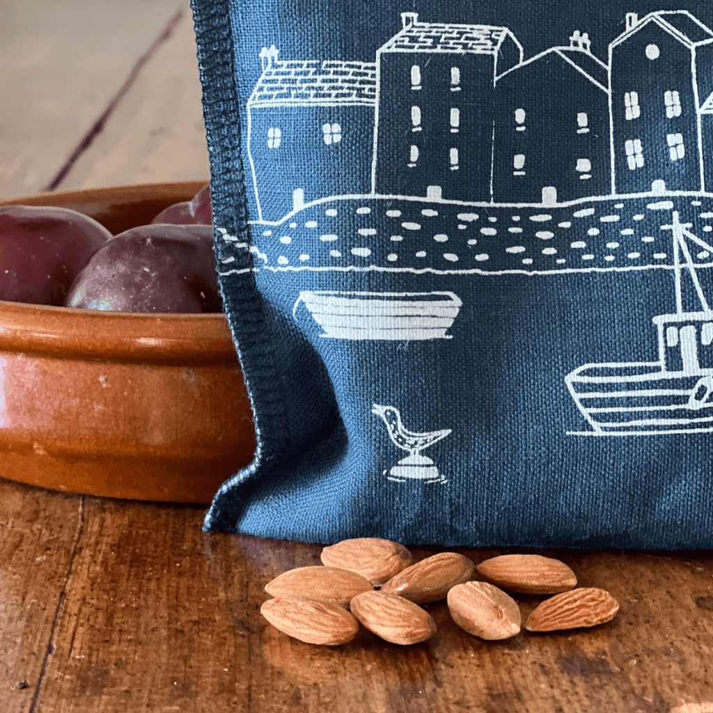 Blue linen snack bag with white house and boat pattern on a wooden surface with almonds and a bowl from Helen Round.