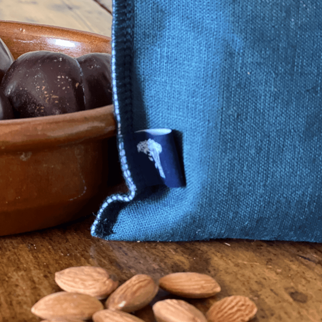 Blue linen snack bag with a white logo on a wooden surface next to almonds and a terracotta bowl from Helen Round.