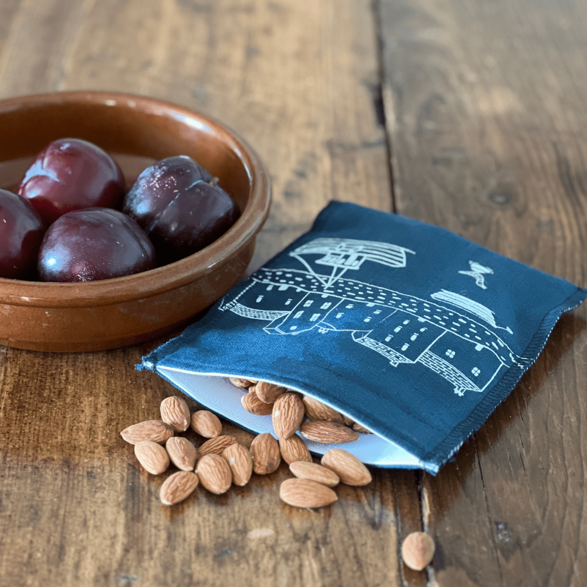 Bowl of plums, almonds, and a blue linen snack bag with white patterns on a wooden surface from Helen Round.