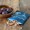 Bowl of plums, almonds, and a blue linen snack bag with white patterns on a wooden surface from Helen Round.