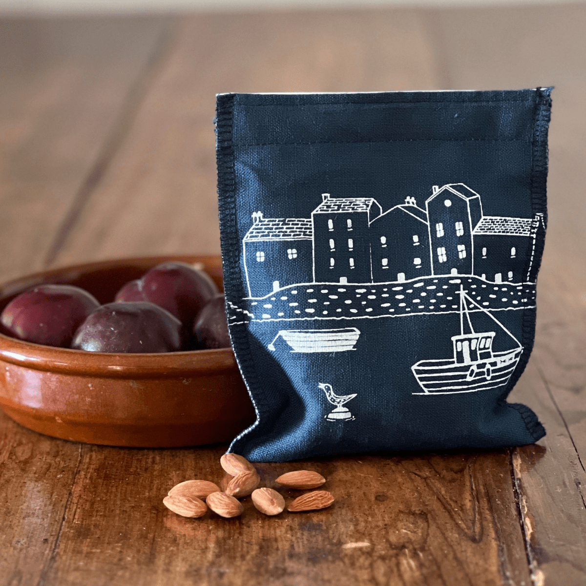 Navy blue linen snack bag with white illustrations of a village and boat on a wooden surface with plums and almonds from the Coastal Collection by Helen Round.