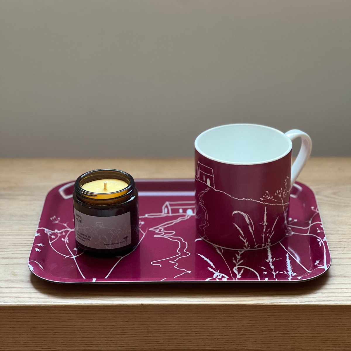 Helen Round bone china mug, Rame Head candle and Rame Head Tray all with an illustrations of Rame Head in white on a raspberry background.