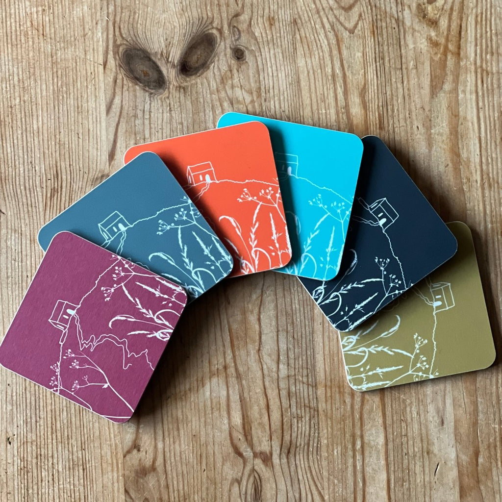 Six colourful coasters featuring the Rame Head design from Helen Round fanned out of a wooden surface.