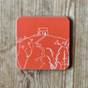 Orange coaster with white Rame Head design, placed on a wooden surface from Helen Round.