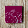 Raspberry red coaster with white Rame Head design placed on a wooden surface from Helen Round.