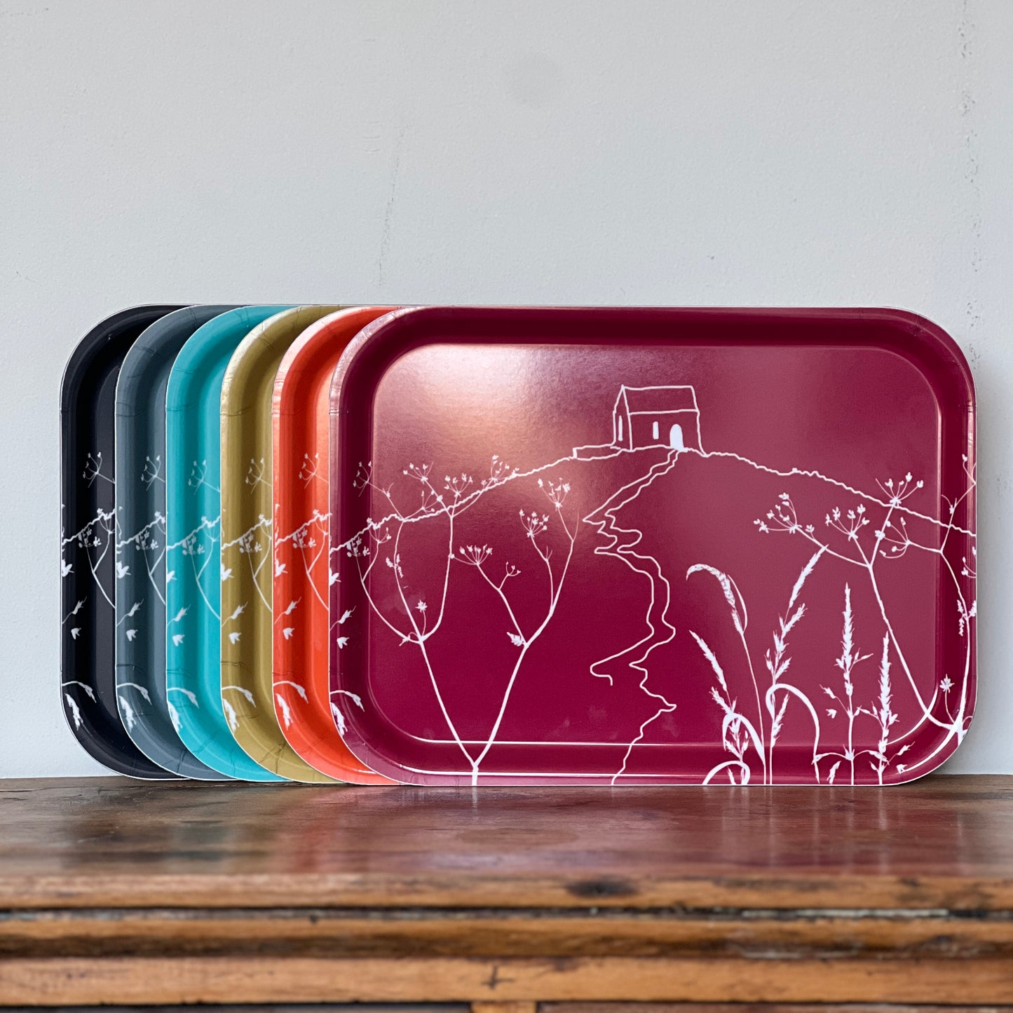 A set of six rectangular trays with rounded corners, fanned out on a wooden surface against a light gray wall. The front tray is  Raspberry with a white line-art illustration of Rame Head, while the trays behind it show additional color options including black, Slate grey, Blue, Green and Orange From Helen Round