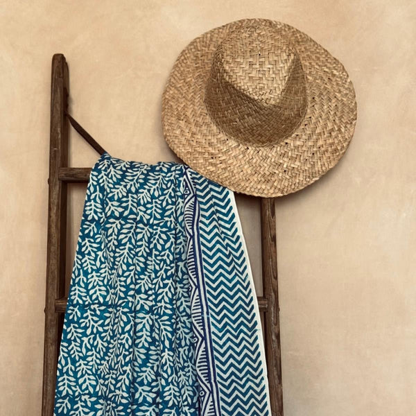 Straw hat and blue hand-printed sarong hanging on a wooden ladder against a beige wall.