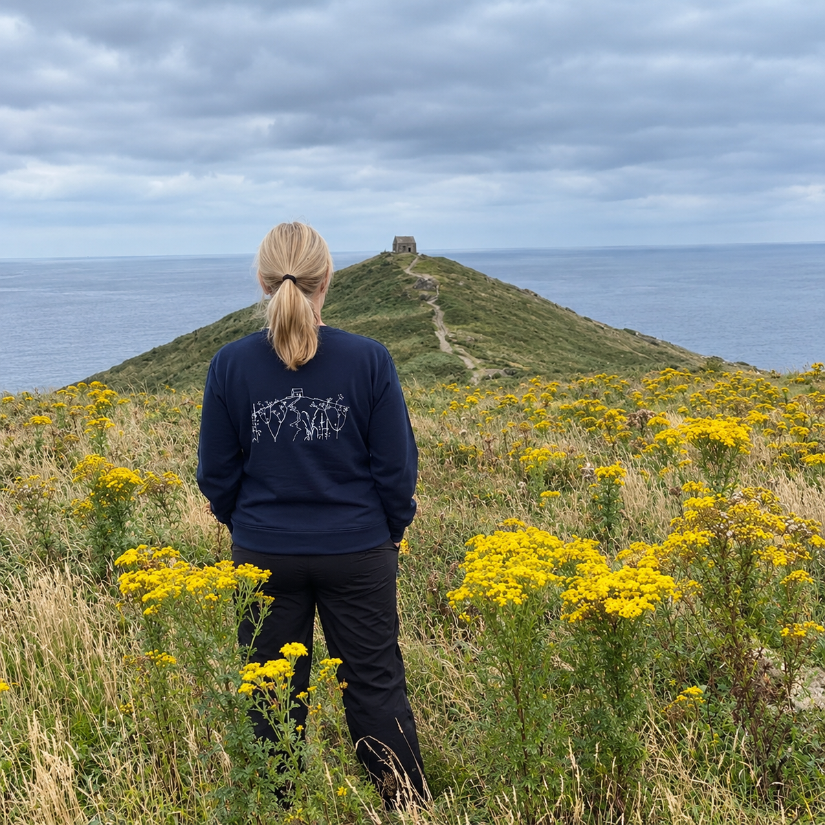 A woman stands at the Rame Head chapel overlooking the ocean, seen from behind. She is wearing a navy blue long-sleeve shirt with a white graphic on the of Rame ead sold by Helen Round