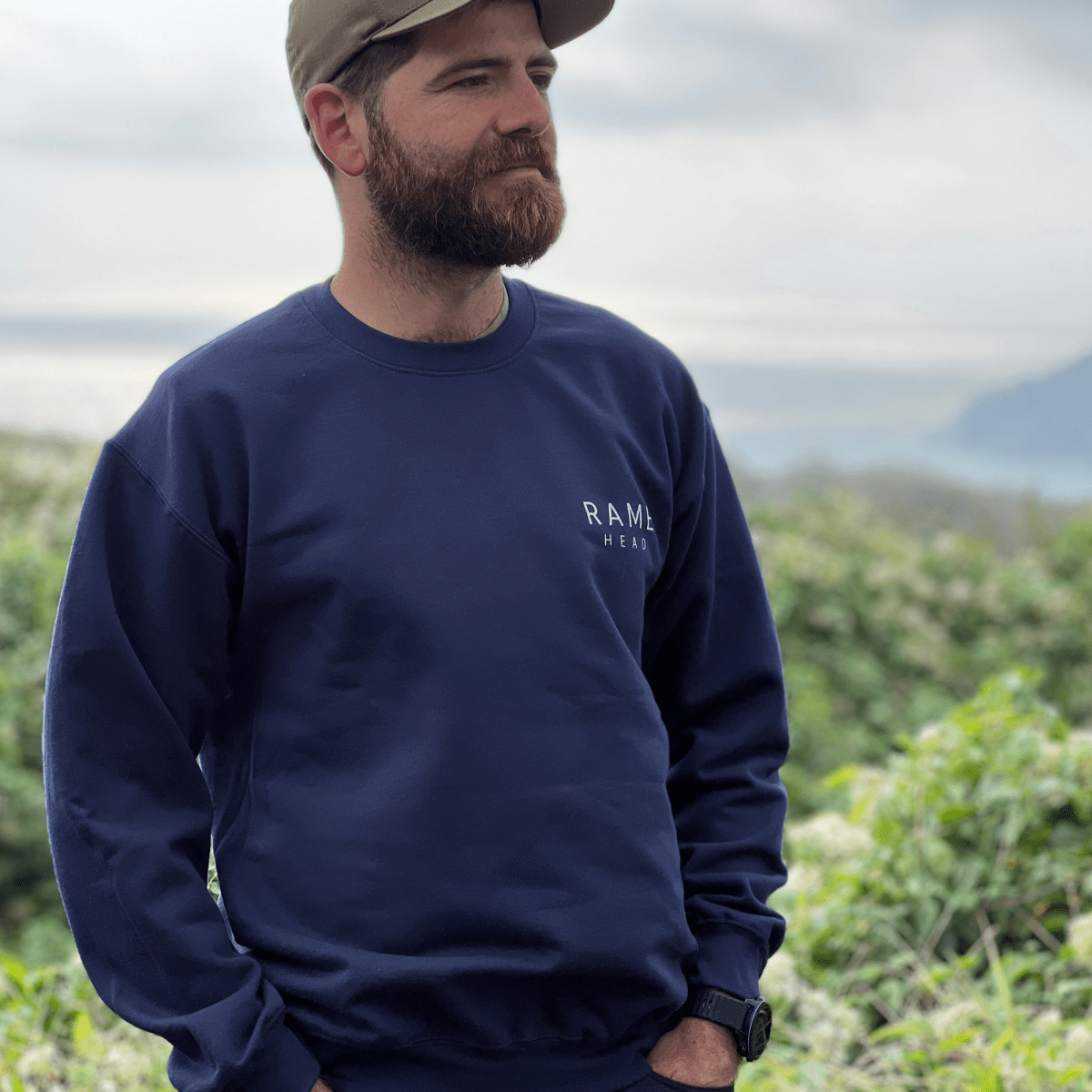 A man wearing a navy blue crewneck sweatshirt with a small white 'RAME HEAD' logo on the chest, paired with a tan baseball cap, standing outdoors against a blurred natural background.