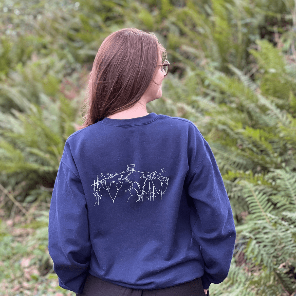 A woman viewed from behind, wearing a navy blue sweatshirt with white line art of Rame Head, set against a background of green ferns from Helen Round