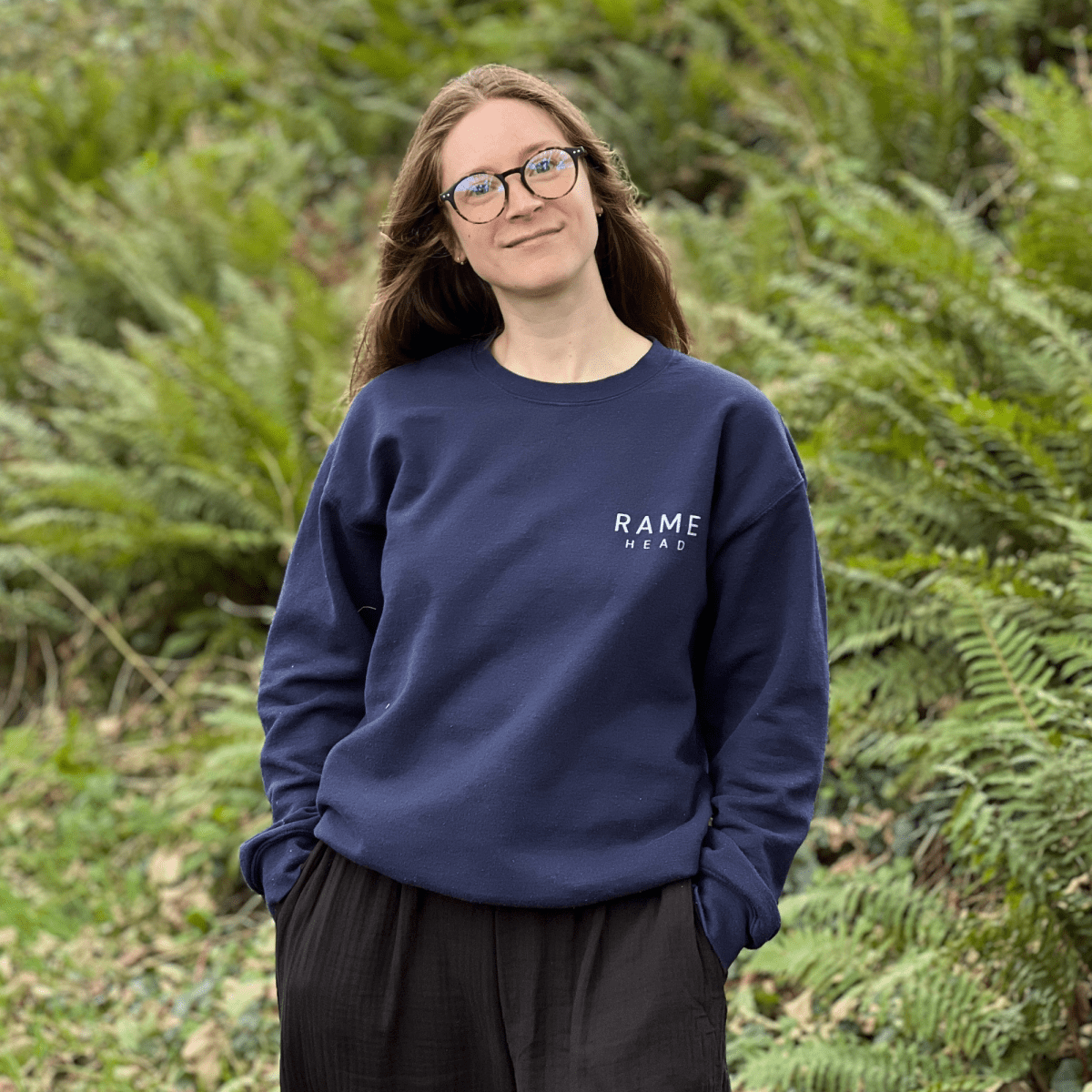 A woman stands outdoors in front of green ferns, wearing a navy blue crewneck sweatshirt with 'RAME HEAD' printed in white on the chest With an illustration of Rame Head on the back from Helen Round
