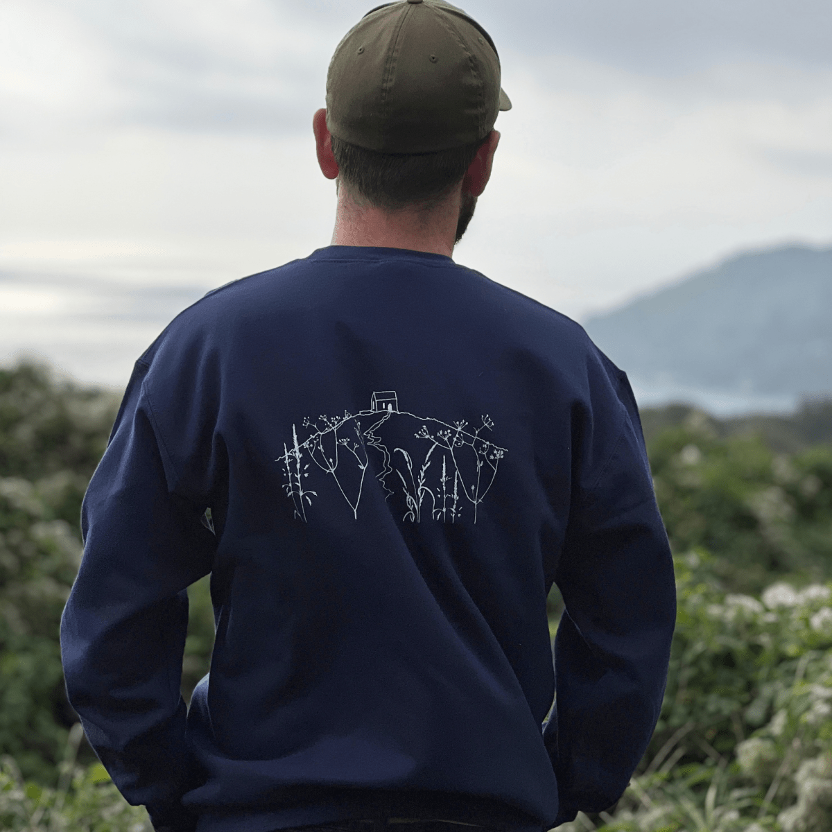 A man viewed from behind wearing a navy blue crewneck sweatshirt with a white line-art rame head on the back, paired with an olive green baseball cap, set against an outdoor background of greenery.